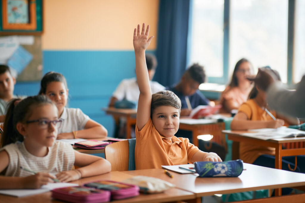 Smiling schoolboy raises his arm to answer teacher’s question during class in classroom.